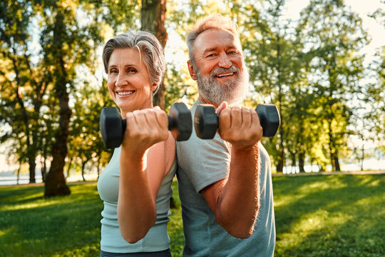 Joint workout on retirement. Beautiful and healthy elderly couple keeping dumbbells in hands while standing back to back at sunny park. Smiling family of two doing exercises for arms on fresh air. - Powered by Adobe