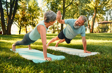 Teamwork during outdoors activity. Strong elderly couple in sportswear standing in plank position, giving high five, looking at each other and smiling. Concept of healthy retirement and active hobby.