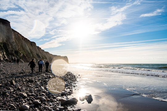 The photograph depicts a group of people walking along a rocky stretch of a beach with towering cliffs on one side. The morning sun shines brightly, creating a flare effect and casting the walkers