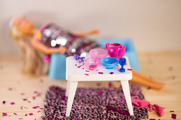 Tired blond plastic doll dressed in sequin dress laying on sofa among chaos after party. Served table, scattered heels and spilled confetti at home