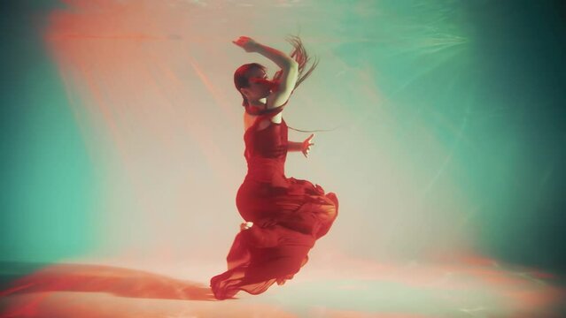 Sexy girl fashion model posing dancing underwater dive creative video shooting waving long legs hands. Fantasy woman swims in dark blue water neon light. silk long red dress. aqua studio pool art 4k