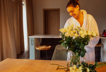Young charming woman in white bathrobe cuts stems of white roses and puts them in glass vase.