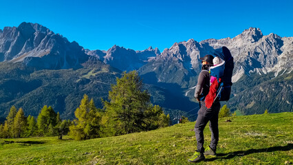 Naklejka na ściany i meble Hiker man with baby carrier with panoramic view of majestic mountain peaks of untamed Sexten Dolomites, South Tyrol, Italy, Europe. Hiking concept in Italian Alps. Looking at mount Dreischusterspitze