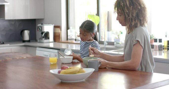 Biracial mother and daughter eating breakfast cereal in sunny kitchen, copy space, slow motion