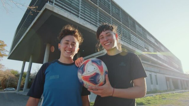 Two happy girls in sportswear standing outdoors on city street, holding soccer ball and posing together on camera with a smile. Zoom shot, video portrait