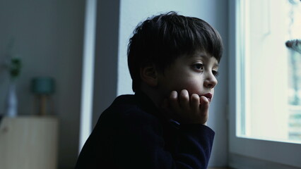 Thoughtful bored child standing at home with hand in chin observing view from apartment window with...