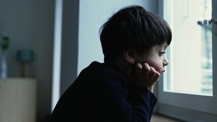 Thoughtful bored child standing at home with hand in chin observing view from apartment window with...