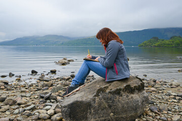 Journalling in the sunshine, pretty young woman sits writing down her thoughts whilst enjoying the peace and quiet of sitting by a lock in rural Scotland. 
