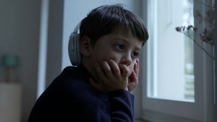 Thoughtful small boy with hand in chin daydreaming at home by window. Pensive child thinking deeply...