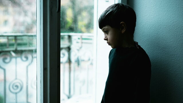 One young boy struggles with mental illness at home with green tint color. Child depressed standing by window with blank expression depicting a meaningless life