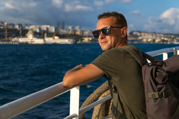 Happy handsome young male tourist in sunglasses with a backpack on his shoulders enjoys the sea and the city from a ferry in Istanbul on a sunny day. Concept of travel, tourism