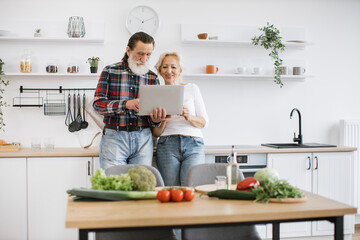 Romantic elderly husband and wife searching internet for recipe for healthy breakfast dish using a modern laptop in bright, spacious kitchen.