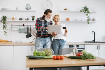 Old Caucasian couple using laptop and credit card to buy food products to prepare breakfast in modern spacious kitchen background.