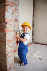 Full length of happy child construction worker holding hammer tool and looking at camera while standing by brick wall at home during renovation. Smiling kid wearing safety helmet and work overalls.