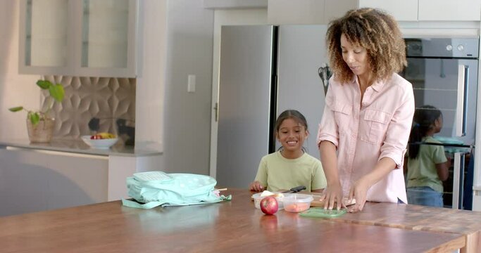 Happy biracial mother and daughter preparing school packed lunch in kitchen, copy space, slow motion