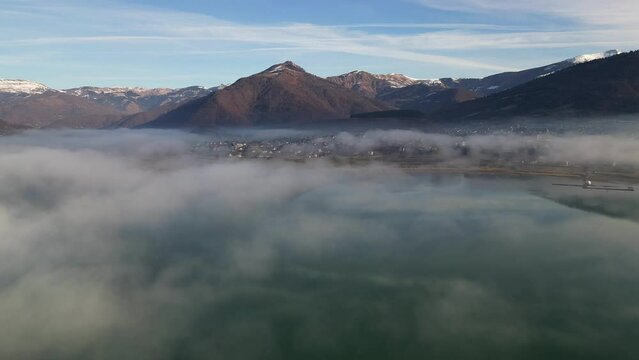 A drone flies over a lake covered in fog, with a mountain village ahead