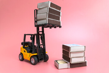A forklift truck holds boxes of matches on a platform and loads them onto a stack from below. The concePink background. Copy space.