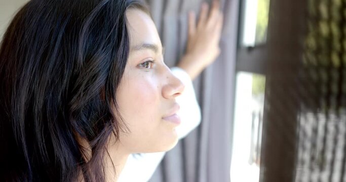 Happy biracial teenage girl drawing curtains, looking out of sunny window, copy space, slow motion