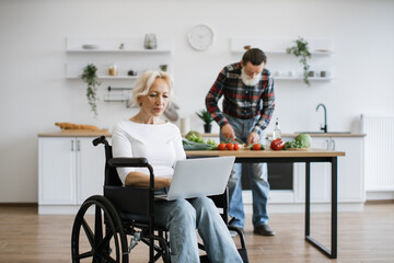 Close up portrait of old woman sits in wheelchair and works on laptop while her husband with gray...