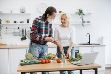 Elderly man with gray beard tears lettuce and herbs into glass bowl while wife cutting peppers in small pieces. Old retired couple preparing fresh healthy salad in modern kitchen.