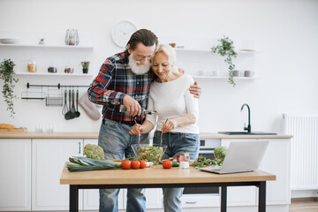 Happy elderly husband and wife preparing healthy salad in bright modern kitchen. Bearded man pours oil in glass bowl while woman stirs pieces of fresh chopped vegetables.