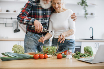 Happy elderly husband and wife preparing healthy salad in bright modern kitchen. Bearded man pours oil in glass bowl while woman stirs pieces of fresh chopped vegetables.