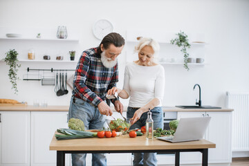 Elderly husband and wife preparing healthy salad in bright modern kitchen. Old man with gray beard mixing pieces of chopped vegetables in glass bowl, while his wife sprinkles salt.
