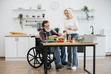 Old couple with disability spends free time cooking breakfast in modern light kitchen. Seniors husband in wheelchair and wife mixing chopped vegetables in bowl while prepare delicious healthy salad. - obrazy, fototapety, plakaty