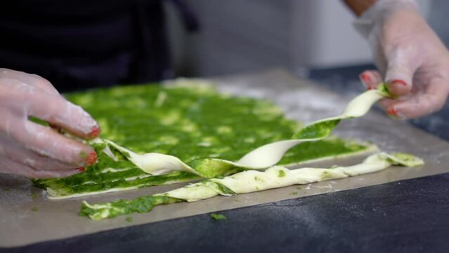 A chef wearing clear gloves rolls ribbons of puff pastry onto gray baking paper. A woman stands at the kitchen table and prepares grissini stuffed with green vegetable paste.