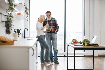 Devoted elderly woman feeds man with gray beard with fresh healthy salad in glass bowl. Happy old...