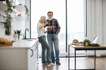 Devoted elderly woman feeds man with gray beard with fresh healthy salad in glass bowl. Happy old pensioner couple having breakfast in modern light kitchen with panoramic windows.