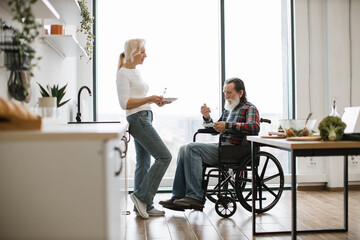 Elderly couple, husband in wheelchair and wife, eating fresh healthy salad from plates with forks in spacious modern bright kitchen with panoramic windows.