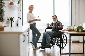 Elderly couple, husband in wheelchair and wife, eating fresh healthy salad from plates with forks...