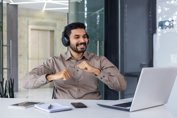 Obraz premium A young Indian man works in the office. He sits at his desk with headphones in front of his laptop and dances during the break, rests, rejoices.