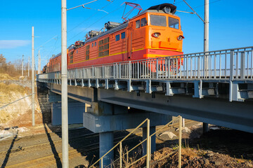 Industrial electric locomotive alternating current pulls opened freight dump rail cars on railroad bridge. AC traction unit.