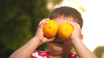 Little son clasps oranges near eyes imitating binoculars spending day in fresh air. Delighted son...