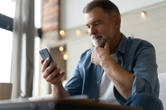 Happy smiling senior man using smartphone device while sitting on sofa at home