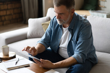 Cheerful senior man in casual clothing using digital tablet while sitting on the sofa at home.