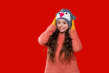 Knitted sweater and beanie hat. Trendy fashion outfit in winter. Winter girl child. Style portrait of teen girl in warm clothing and winter hat isolated on red. Fleece-lined earflaps