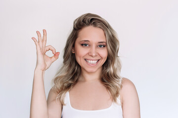 A young caucasian attractive blonde woman with wavy hair showing okay gesture with her hand isolated on a white background. Positive human emotion. The OK approval sign - obrazy, fototapety, plakaty