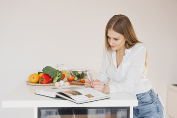 A happy cute blonde woman is writing something down and looking at a recipe book in the kitchen