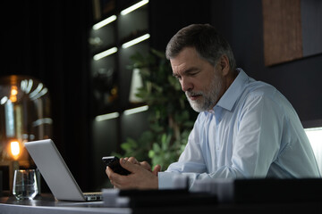 Fototapeta na wymiar Smiling mature businessman holding smartphone sitting in office. Middle aged manager ceo using cell phone mobile apps and laptop. Digital technology applications and solutions for business development