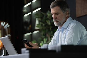 Smiling mature businessman holding smartphone sitting in office. Middle aged manager ceo using cell phone mobile apps and laptop. Digital technology applications and solutions for business development