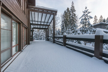 The facade of a wooden house with a spacious terrace against the background of snow-covered fir...