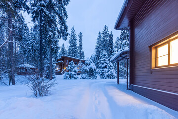 Private territory with snow-covered fir trees. Twilight. Wooden house with bright light .from the window.
