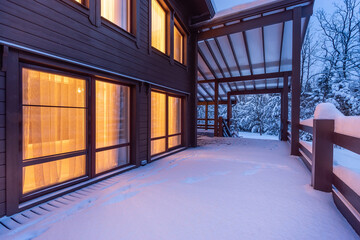 A terrace with a canopy on the background of snow-covered trees. Twilight. Light from the .windows of a wooden house.