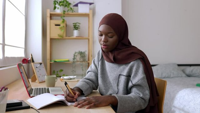 Young adult african student woman taking notes while using laptop computer at home. Millennial black female in muslim headscarf learning online listening virtual video call. High quality 4k footage