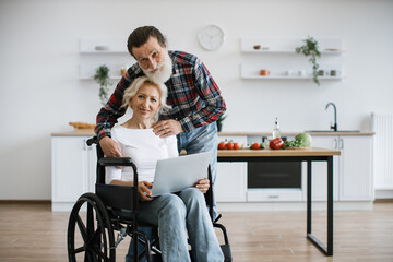Close up of carming old couple of woman in wheelchair with laptop and husband hugging her on background of bright modern kitchen, looking at camera.