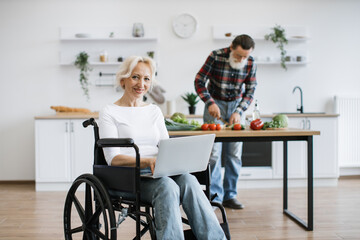 Portrait of old woman looking at camera sits in wheelchair and works on laptop while her husband...