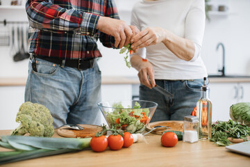 Close up of hands of elderly man and woman in casual clothes tears lettuce and herbs into glass bowl. Old retired couple preparing fresh healthy salad in modern kitchen at home.
